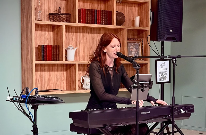 Lollirox chante et joue du piano numérique lors d’une prestation, devant une étagère en bois